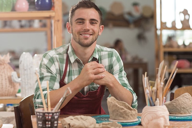 A man with a chiseled jaw wearing a checkered shirts and red apron sits in front of his clay in a workshop, learning how to sculpt. Just like he create beautiful artwork, St. Louis Cosmetic Dentist Dr. Chris Hill is able to sculpt a better jaw with cosmetic dentistry.
